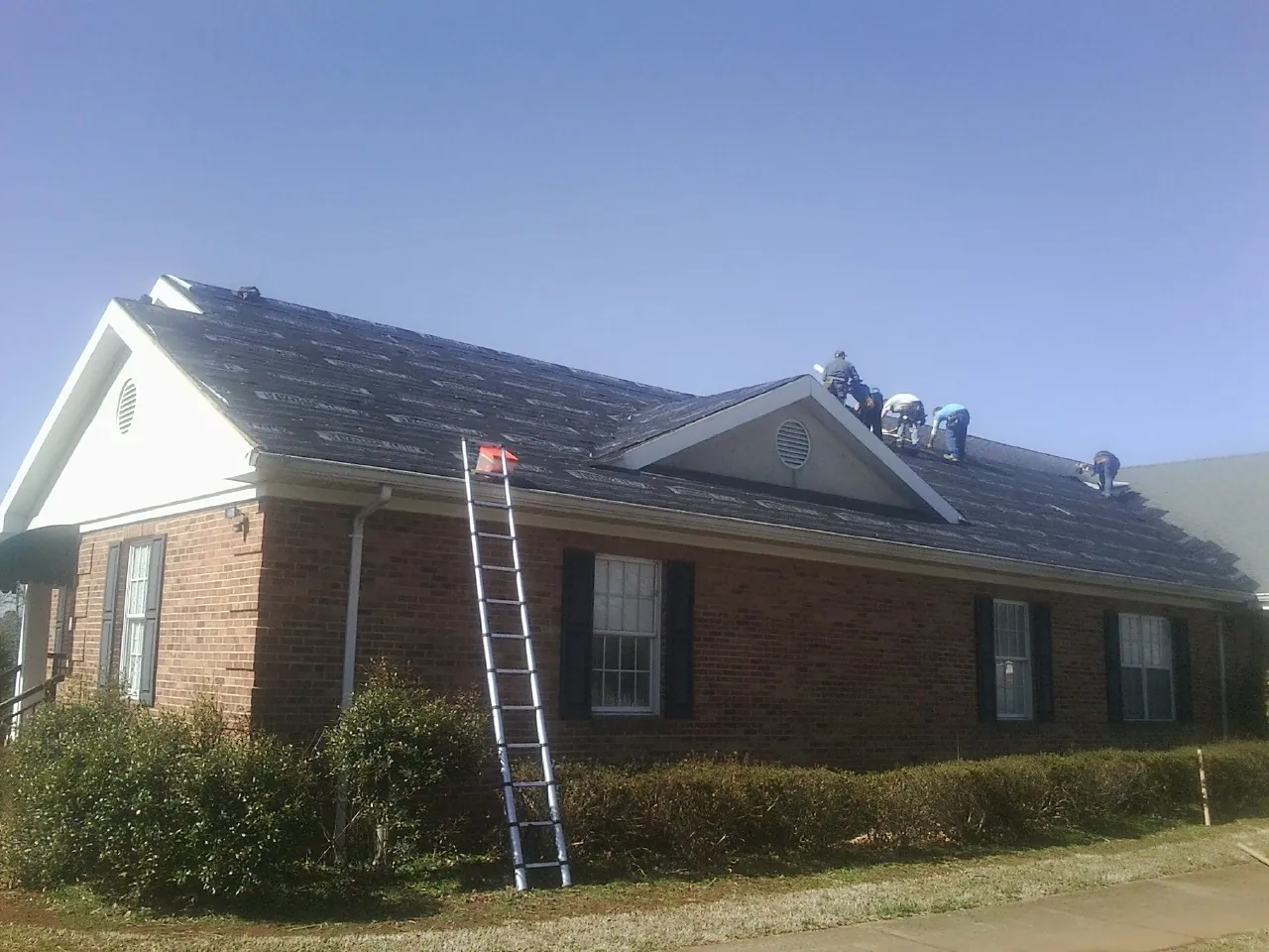 Crew tearing shingles off a brick ranch roof with a ladder set — Metal Roof Repair work by our crew in Cowansville