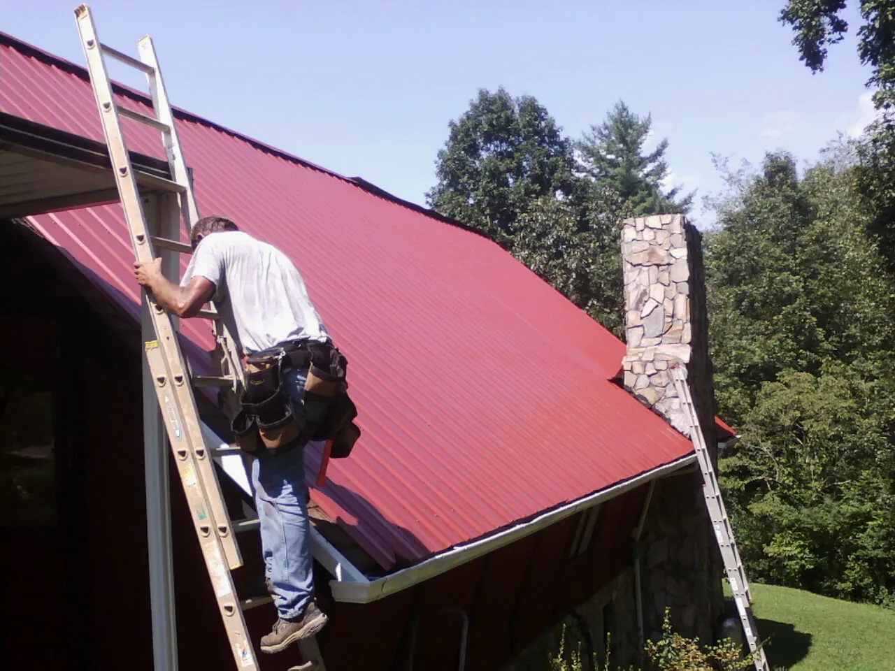 Roofer on a ladder beside a red metal roof and stone chimney — Soffit & Fascia Repair by Roofing Brothers in Cowansville